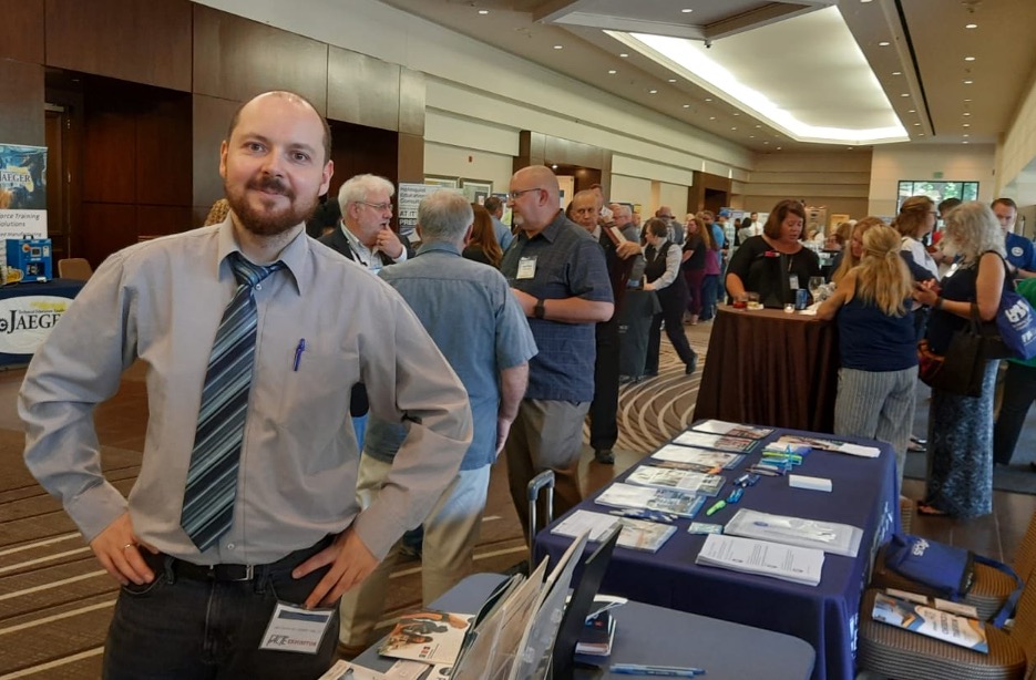 Grzegorz Czuchaj standing with the OpenEDG table at the FACTE conference.